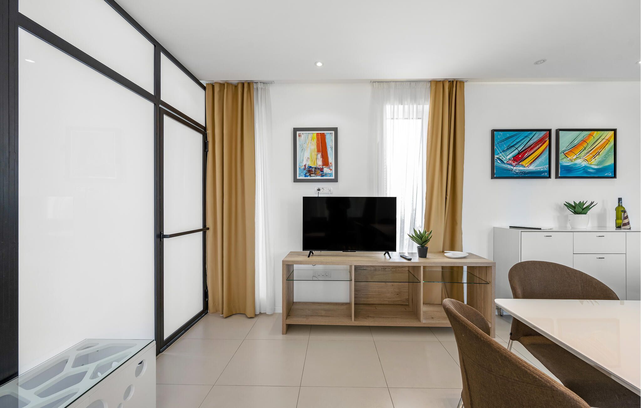 Una sala de estar moderna con un televisor de pantalla plana, una mesa de comedor y un sofá. Hay dos cuadros en la pared y una planta en maceta sobre la mesa.