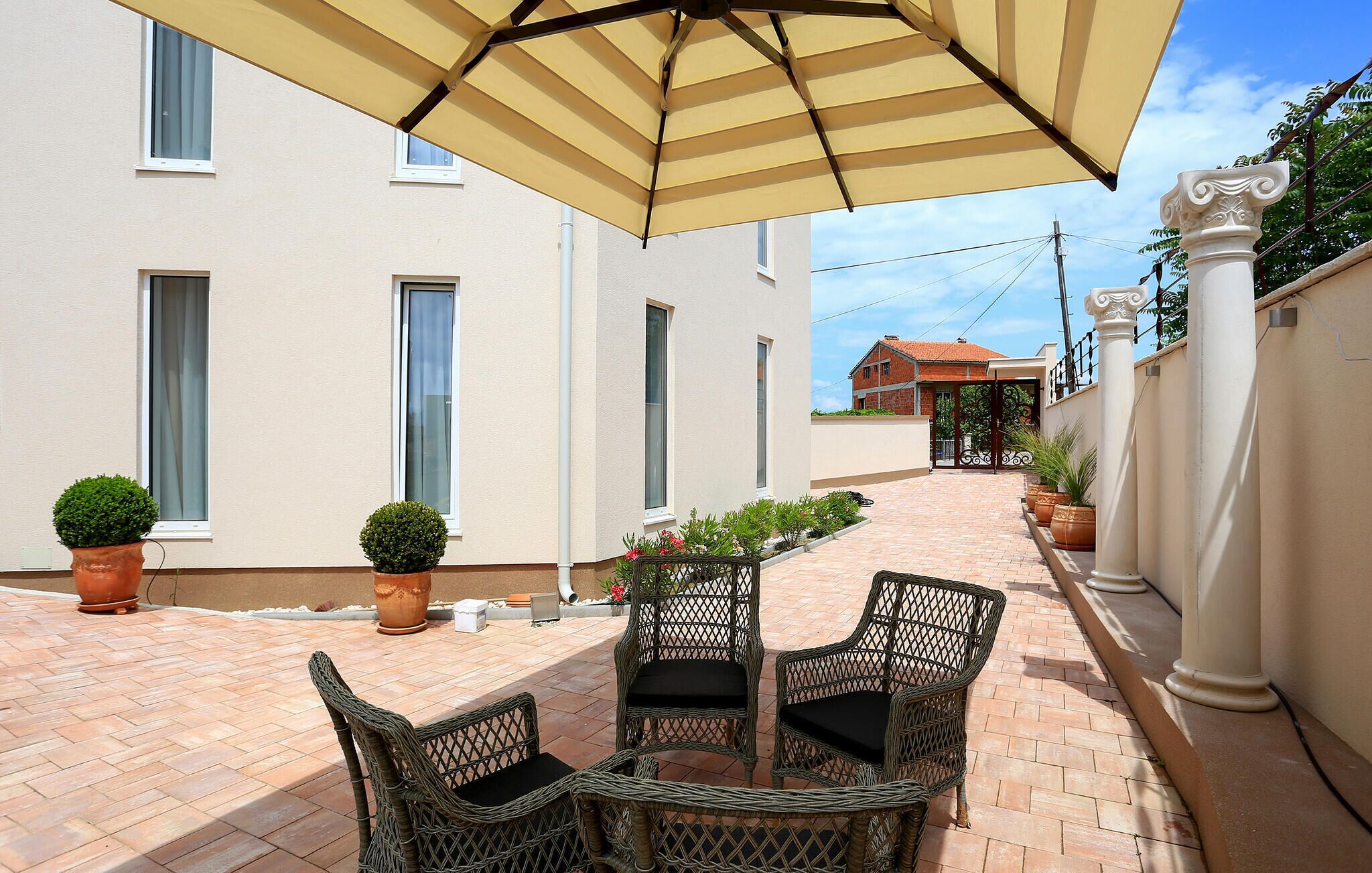 A patio with wicker chairs and a striped umbrella, a tiled pathway, and a building with multiple windows and a gate in the background.