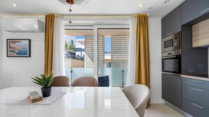 Una cocina moderna con horno empotrado y microondas, un comedor con mesa y sillas blancas, y vistas a la costa a través de amplias ventanas con cortinas amarillas.