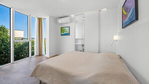 Un dormitorio con una cama grande, una puerta corrediza de vidrio que da a un balcón, un televisor montado y un cuadro en la pared.