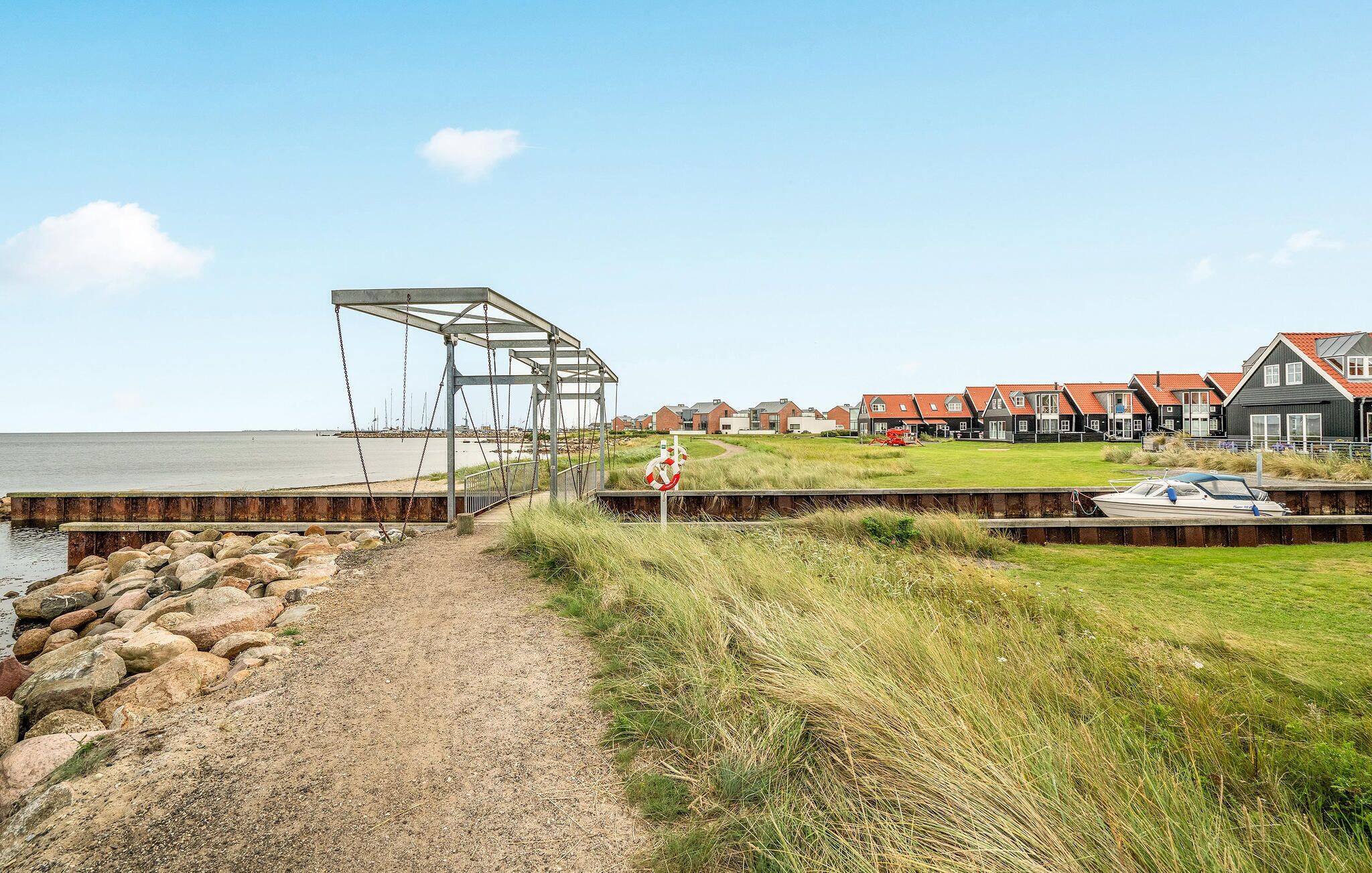 En kystvandring fører til en strand med en metalstruktur, et række huse med røde tage og en båd fortøjet til højre.