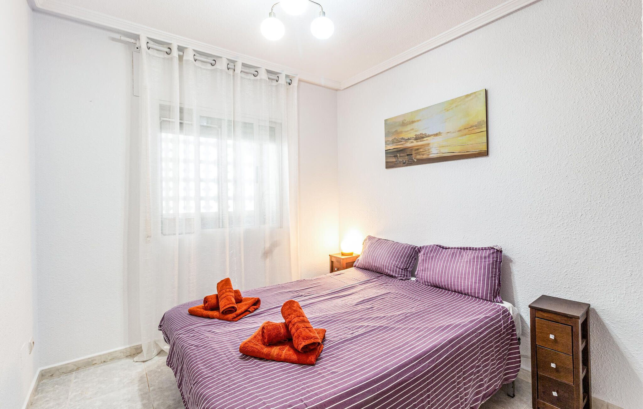 A bedroom with a bed, a nightstand, a lamp, a painting, and a window with curtains.