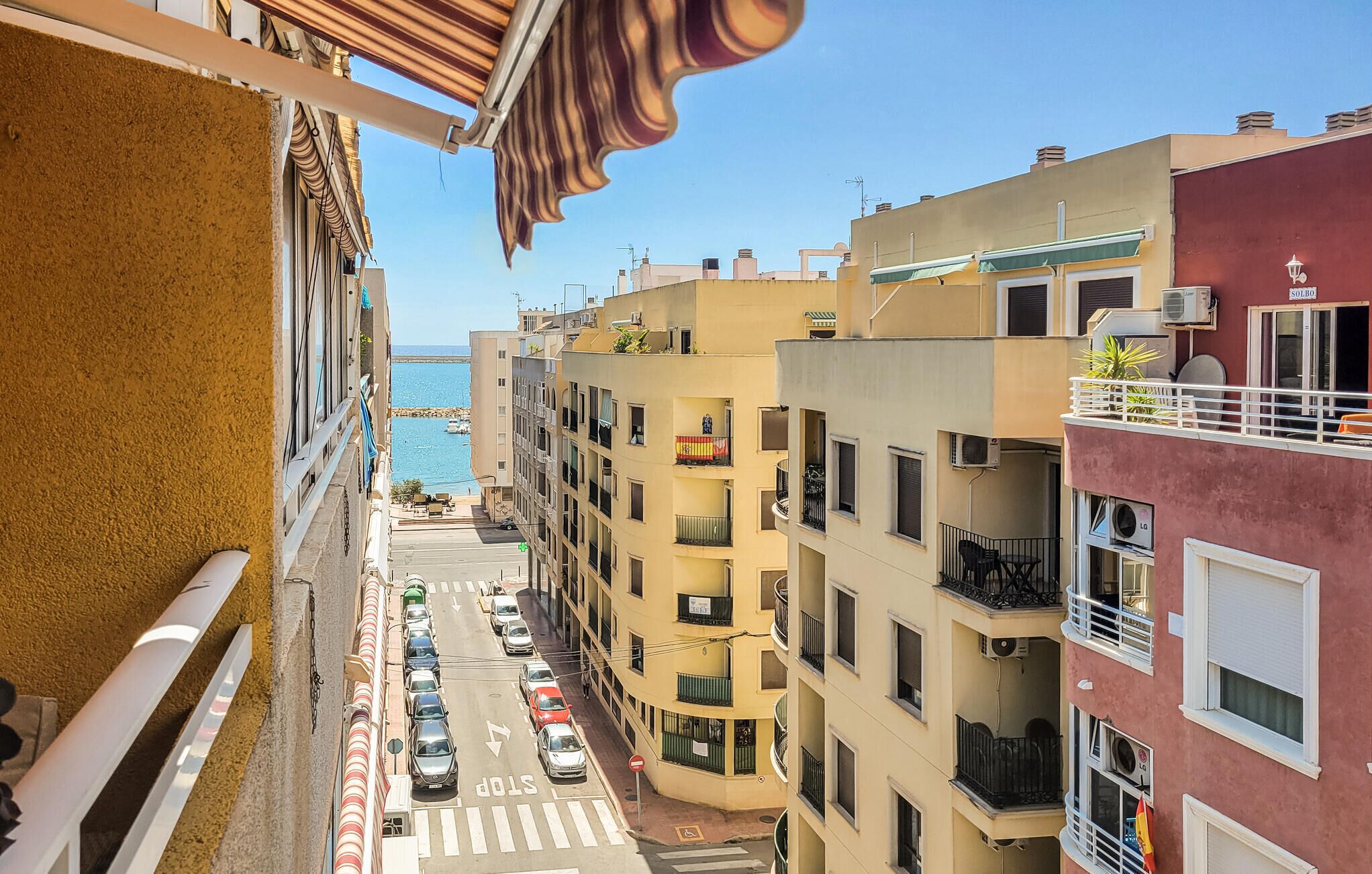 Una calle estrecha bordeada por edificios de varios pisos con balcones, con vista despejada al mar y coches estacionados.