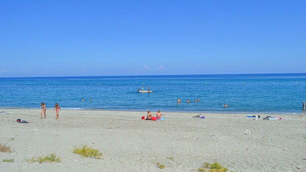 Une plage avec un ciel dégagé, une eau calme, et des gens qui se baignent et prennent des bains de soleil.