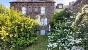A stone building with a garden in front, featuring white shutters and a small door.