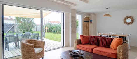 A spacious living area with a large sliding glass door leading to a patio, a wicker armchair, a round coffee table, and a sofa with orange cushions.