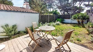 Una terrassa de fusta amb una taula rodona i quatre caires, envoltada per un jardí ple de plantes diverses i una paret blanca al fons.
