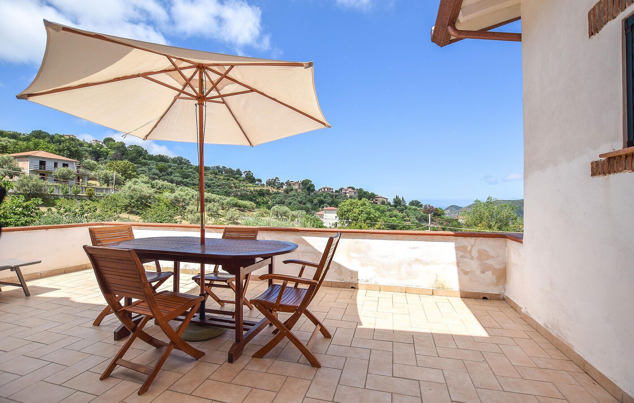 Terraço com mesa e cadeiras, um grande guarda-sol e vista para uma colina com casas.