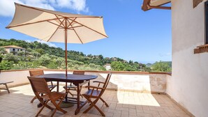 A terrace with a table and chairs, a large umbrella, and a view of a hillside with houses. - Amazing home in Perdifumo (Perdifumo)