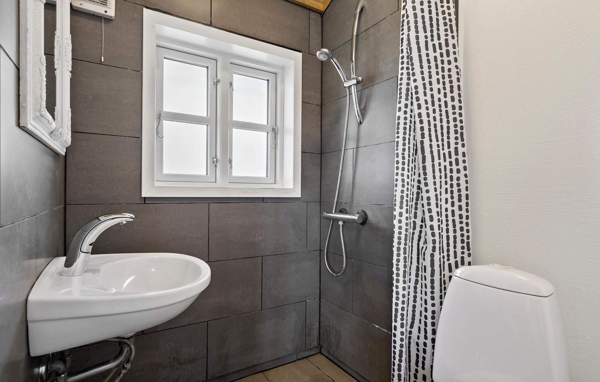 Een badkamer met een witte wastafel, een douche met een handdouche, een toilet en een raam met een witte omlijsting.