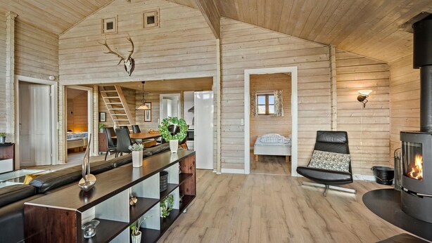 A spacious cabin with a high ceiling, a central dining area with a long table, a cozy seating area with a fireplace, and a visible staircase leading to a loft.