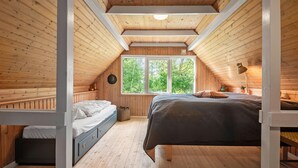 A cozy attic bedroom with a sloped ceiling, wooden walls, a large window, a bed, a nightstand, and a lamp.
