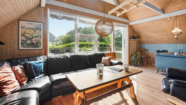 A cozy room with a black leather sofa, a wooden table, and a large window offering a view of greenery.