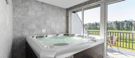 Une salle de bain moderne dotée d’un grand jacuzzi, d’un banc et offrant une vue sur un paysage verdoyant grâce à de grandes portes coulissantes.