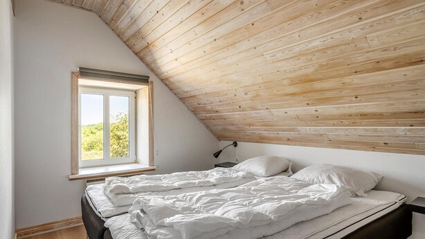 Un dormitorio con techo inclinado, una ventana con vistas a la vegetación, una cama con ropa blanca y una mesita de noche con lámpara.