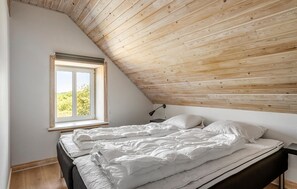 Un dormitorio con techo inclinado, una ventana con vistas a la vegetación, una cama con ropa blanca y una mesita de noche con lámpara.