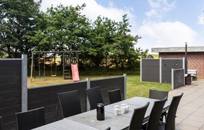 Un patio al aire libre con una mesa y sillas, un parque infantil con un tobogán y un edificio de ladrillo al fondo.