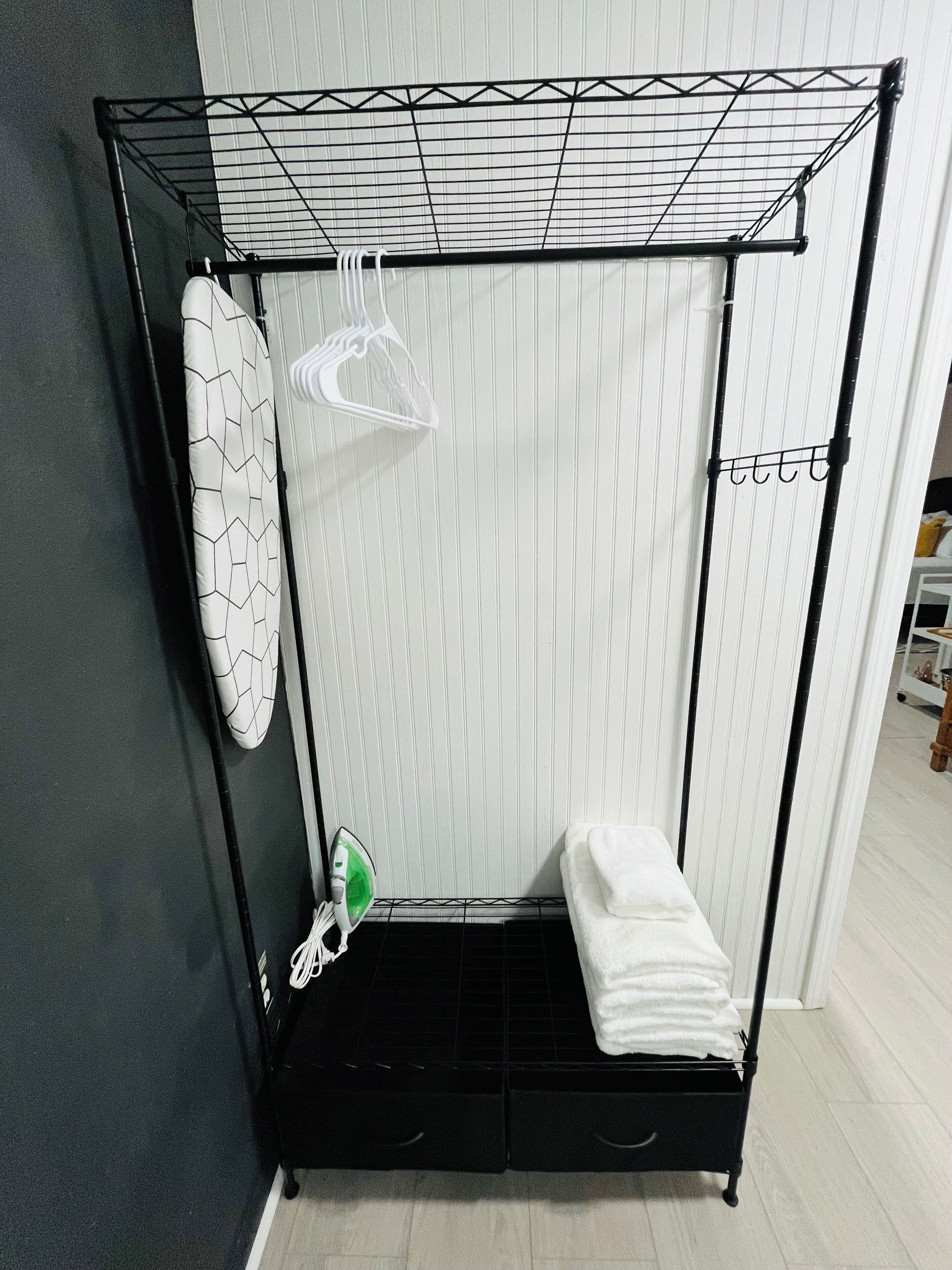 A corner room with a black metal drying rack, a white ironing board, and a white towel on a black shelf.