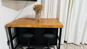 Ett litet träbord med två svarta barstolar, en vas med torkade blommor och en tv som är monterad på väggen.