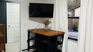 Ett rum med ett träbord, två svarta barstolar, en väggmonterad tv och ett fönster med vita gardiner.