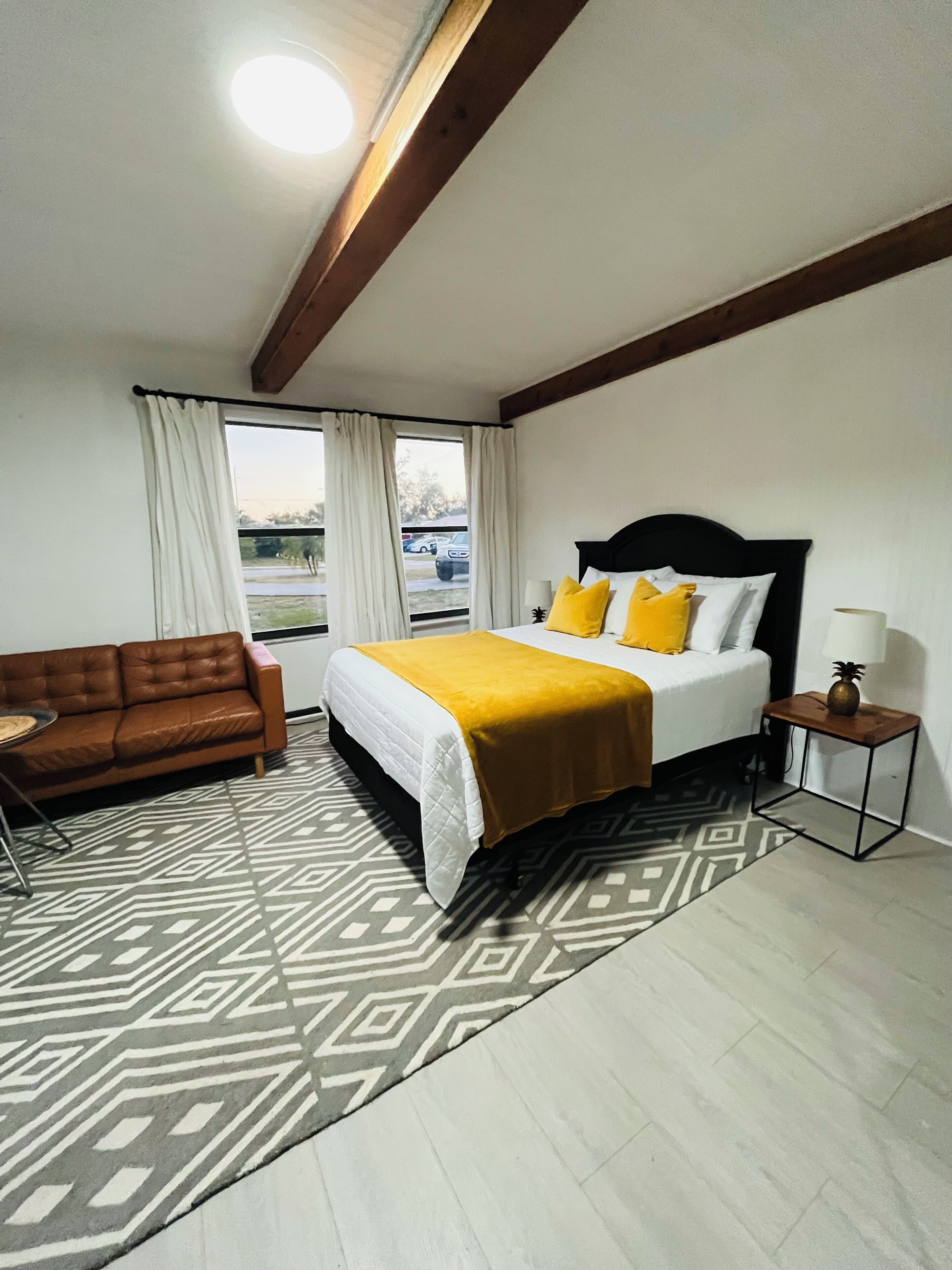 A hotel room with a bed, a brown leather sofa, a patterned rug, and a window with curtains.