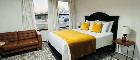 A hotel room with a bed, a brown leather sofa, a patterned rug, and a window with curtains.