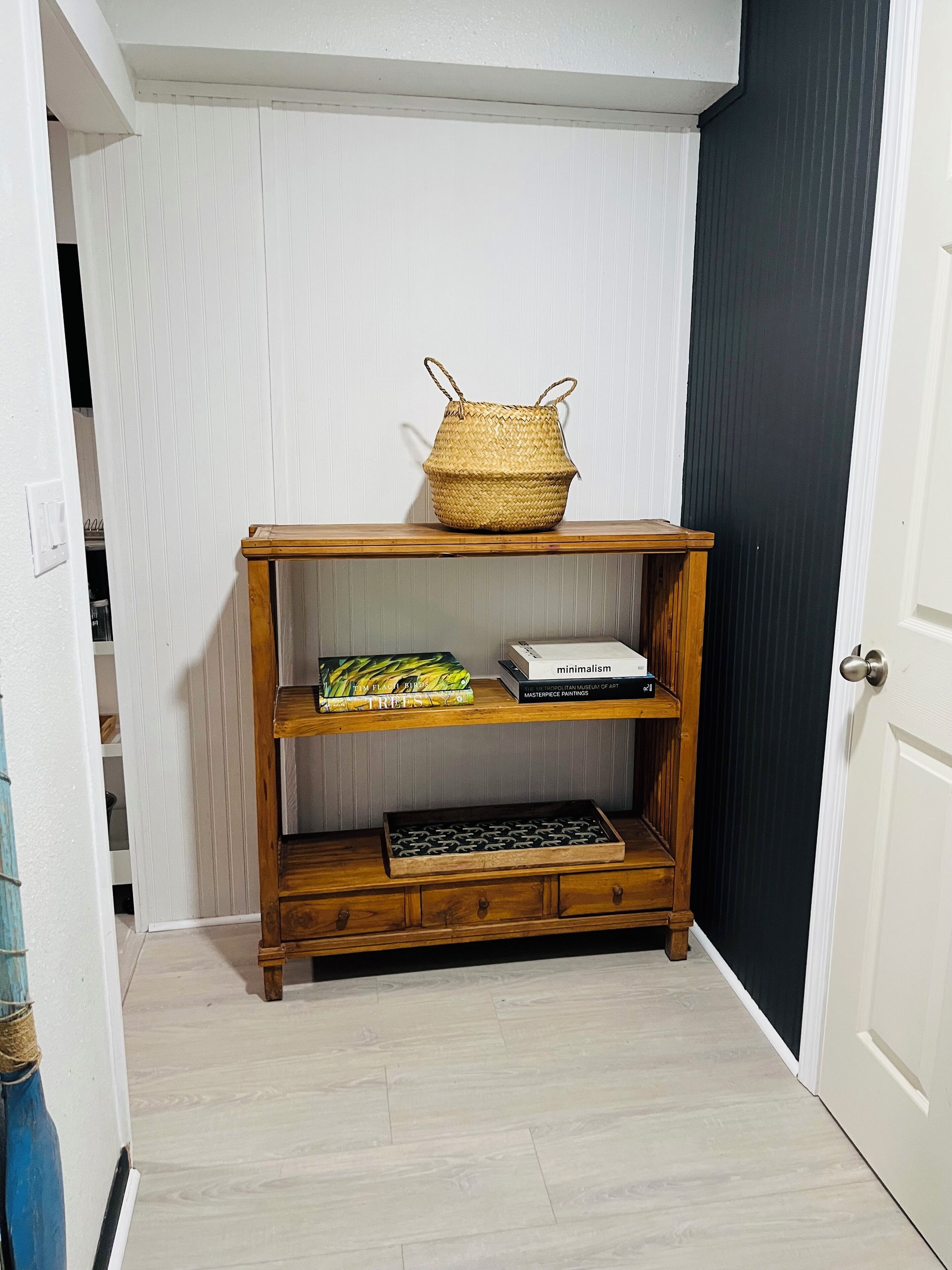 A wooden shelf with a woven basket on top, a book titled 'minimalism', and a box labeled 'MASTERCHEF PAINTINGS'.