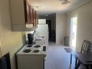 A compact kitchen with a stove, a coffee maker, and a refrigerator. There is a dining area with a table and chairs, and a trash can near the window. - Charming 1-bedroom tiny house in awesome Chester with WiFi, central AC and Heat. (Chester)