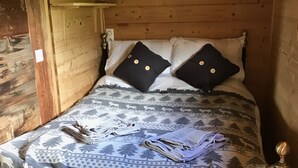Dormitorio de cabaña de madera con cama, dos almohadas, lámpara y manta con estampado de bosque y ciervos.
