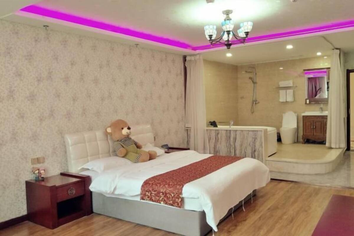 Habitación de hotel con cama, mesita de noche, un oso de peluche, baño con ducha y espejo.