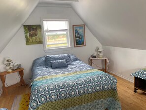 A bedroom with a sloped ceiling, a bed with patterned bedding, a window with blinds, a bedside table with a lamp, and a small table with a tissue box.