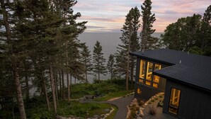 Una casa moderna con un sendero de piedra que lleva a una terraza de madera, rodeada de un bosque denso y con vistas a un cuerpo de agua.