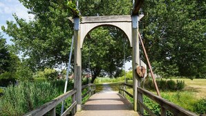 Un puente peatonal de madera con diseño en arco, rodeado de abundante vegetación y una masa de agua tranquila.