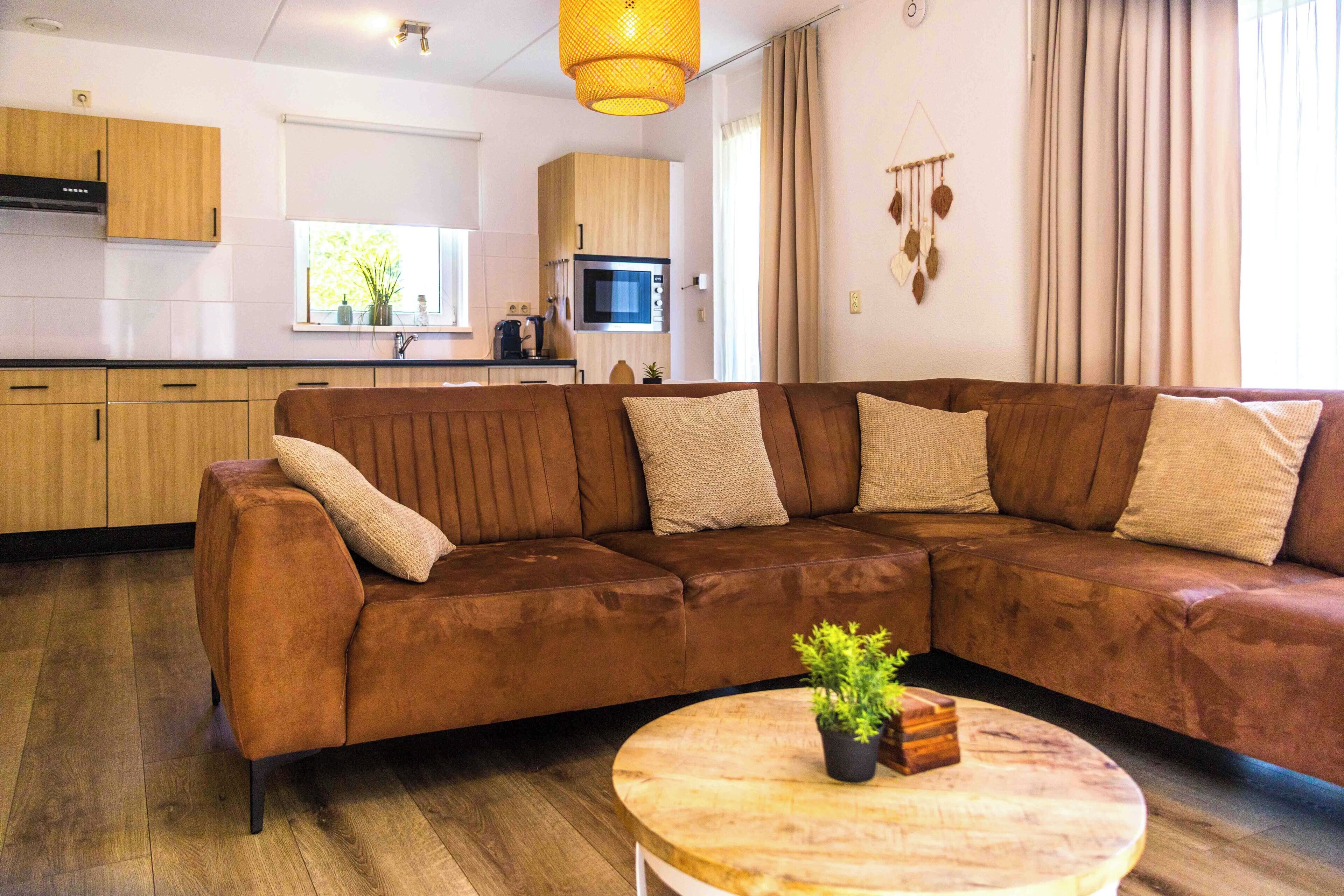 Ein modernes Wohnzimmer mit einem braunen Ecksofa, einem runden Couchtisch aus Holz und einer Küche im Hintergrund.