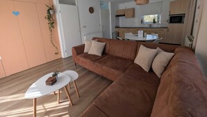 Una sala moderna con un sofá seccional marrón, una mesita blanca y una cocina al fondo.