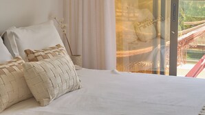 Ein Schlafzimmer mit einem großen Bett, weißer Bettwäsche, einer gewobenen Lampe und Blick auf eine Hängematte sowie Palmen durch eine Glastür.