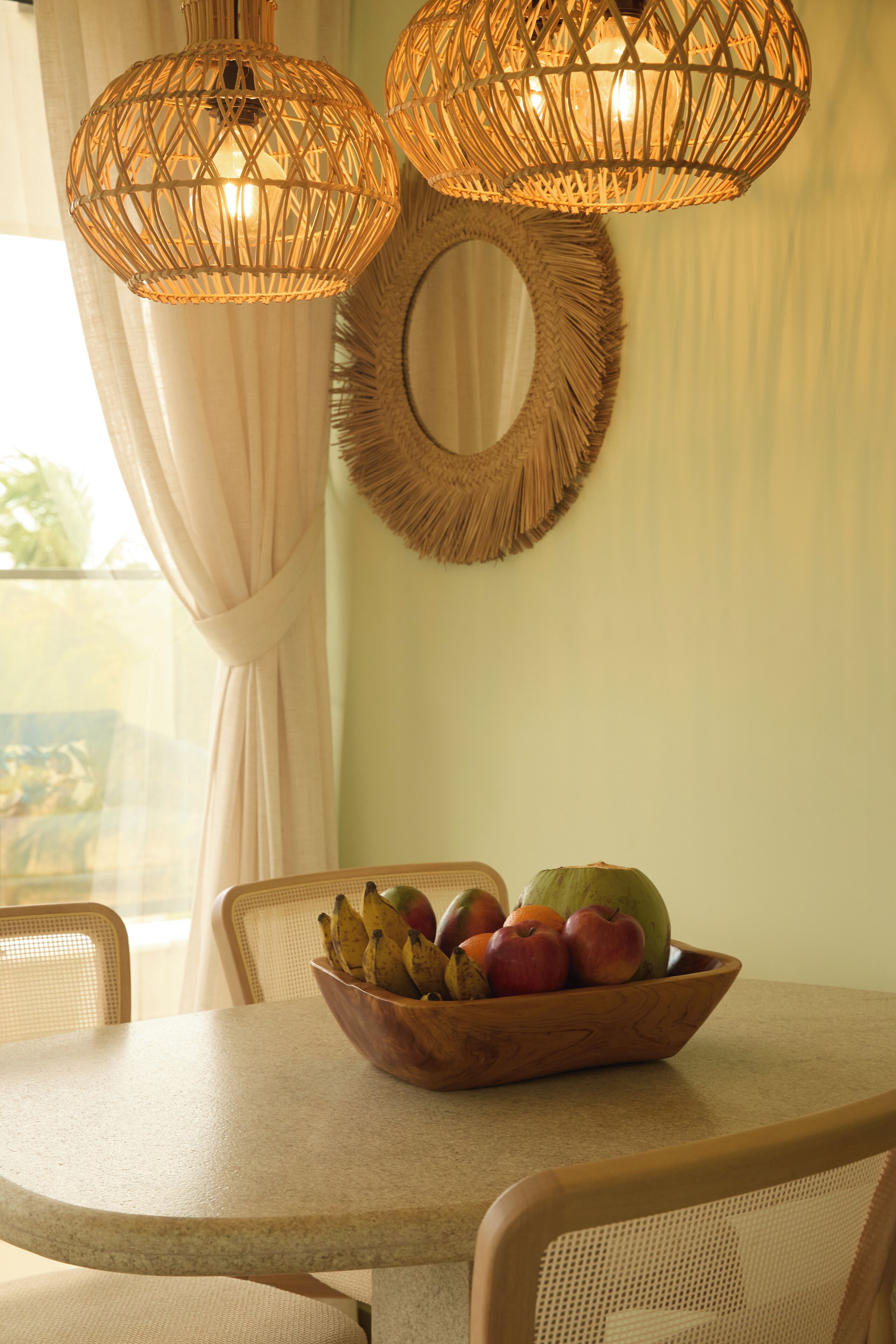 A dining area with a round table, wicker chairs, a fruit bowl, and two wicker pendant lights.