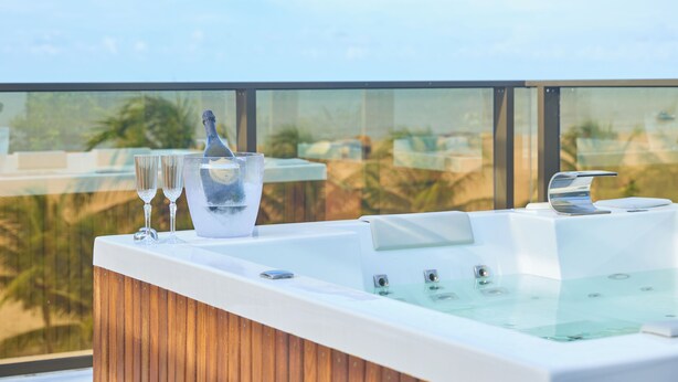 Ein moderner Whirlpool mit Holzvertäfelung, zwei Gläsern und einer Flasche auf einem Balkon mit freiem Blick auf Palmen und blauen Himmel.