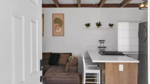 Et kompakt køkken med en hvid bordplade, indbygget komfur og en lille ø. I det tilstødende opholdsområde er der en brun sofa og en hvid stol.