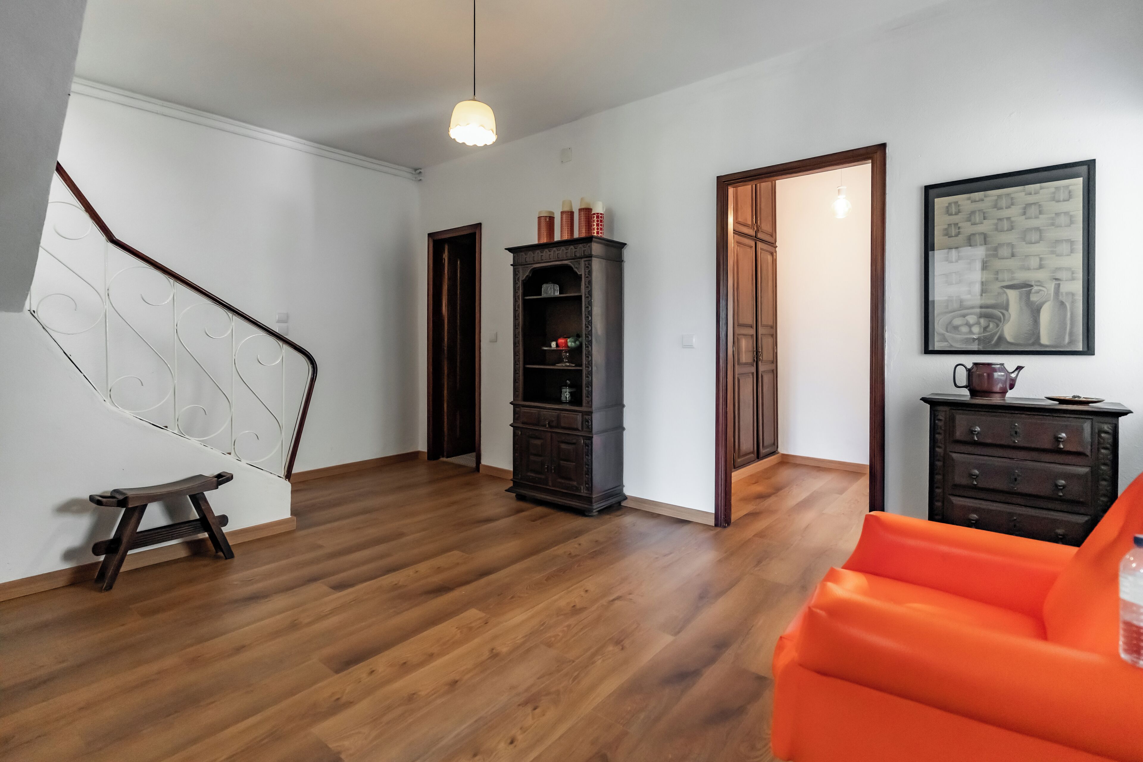Une pièce spacieuse avec un plancher en bois, un escalier, un meuble en bois foncé et une chaise orange vif.