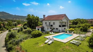 Una casa de dos pisos con piscina, rodeada de abundante vegetación y un cielo azul despejado.