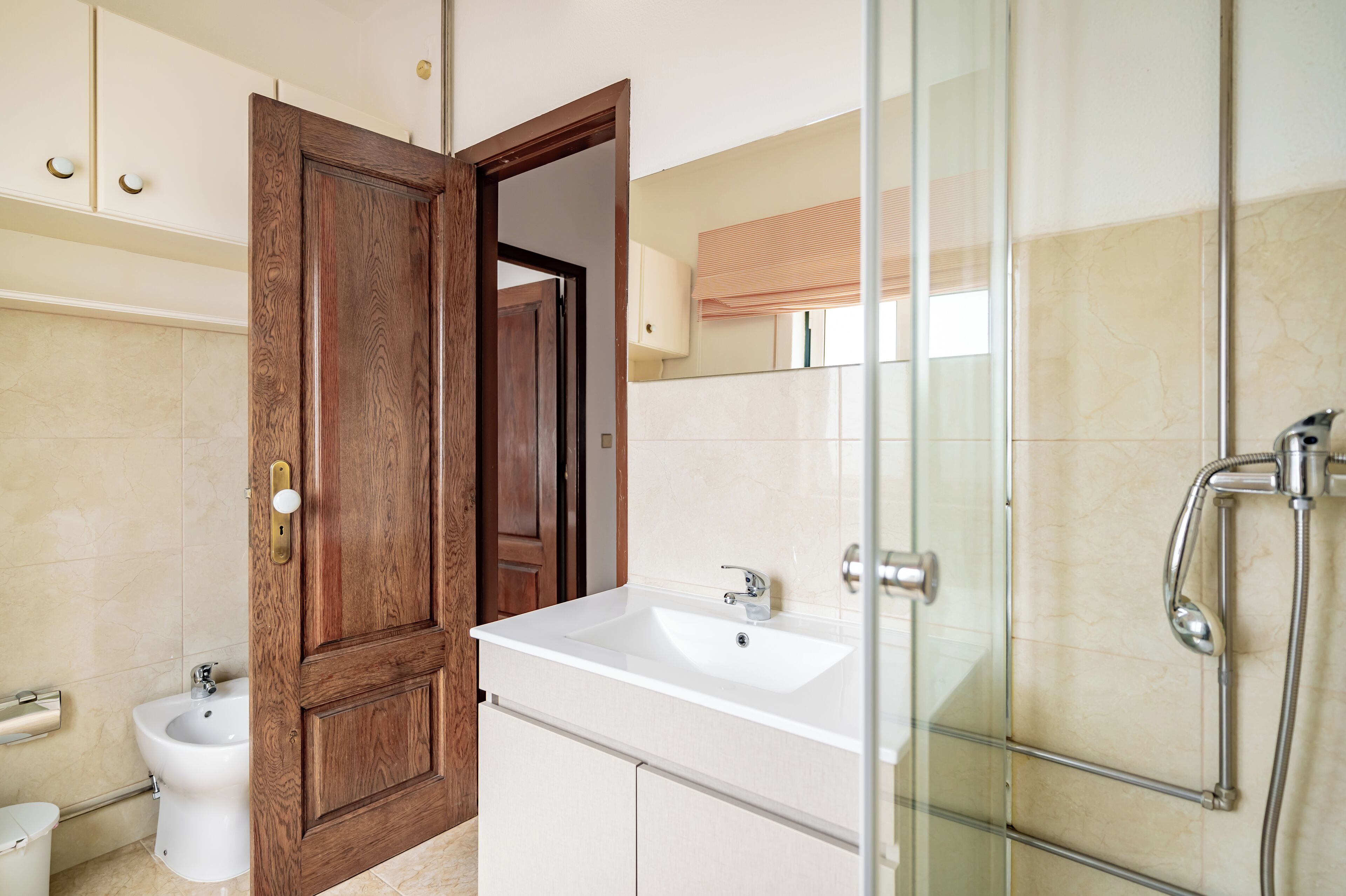 Une salle de bain avec une porte en bois, un lavabo blanc, un bidet et une cabine de douche en verre.