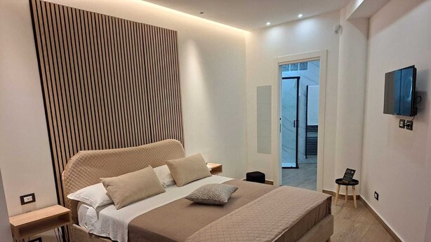 A modern bedroom with a large bed, a bedside table, a TV mounted on the wall, and a view of the outside through a door.