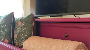 Ein pinkfarbenes Bett mit einer gesteppten Decke, darüber ein Fernseher und ein Kissen mit Blumenmuster.
