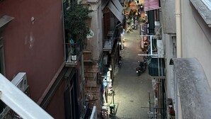 Une rue étroite en milieu urbain, bordée d’immeubles à plusieurs étages dotés de balcons, dont certains sont ornés de plantes.