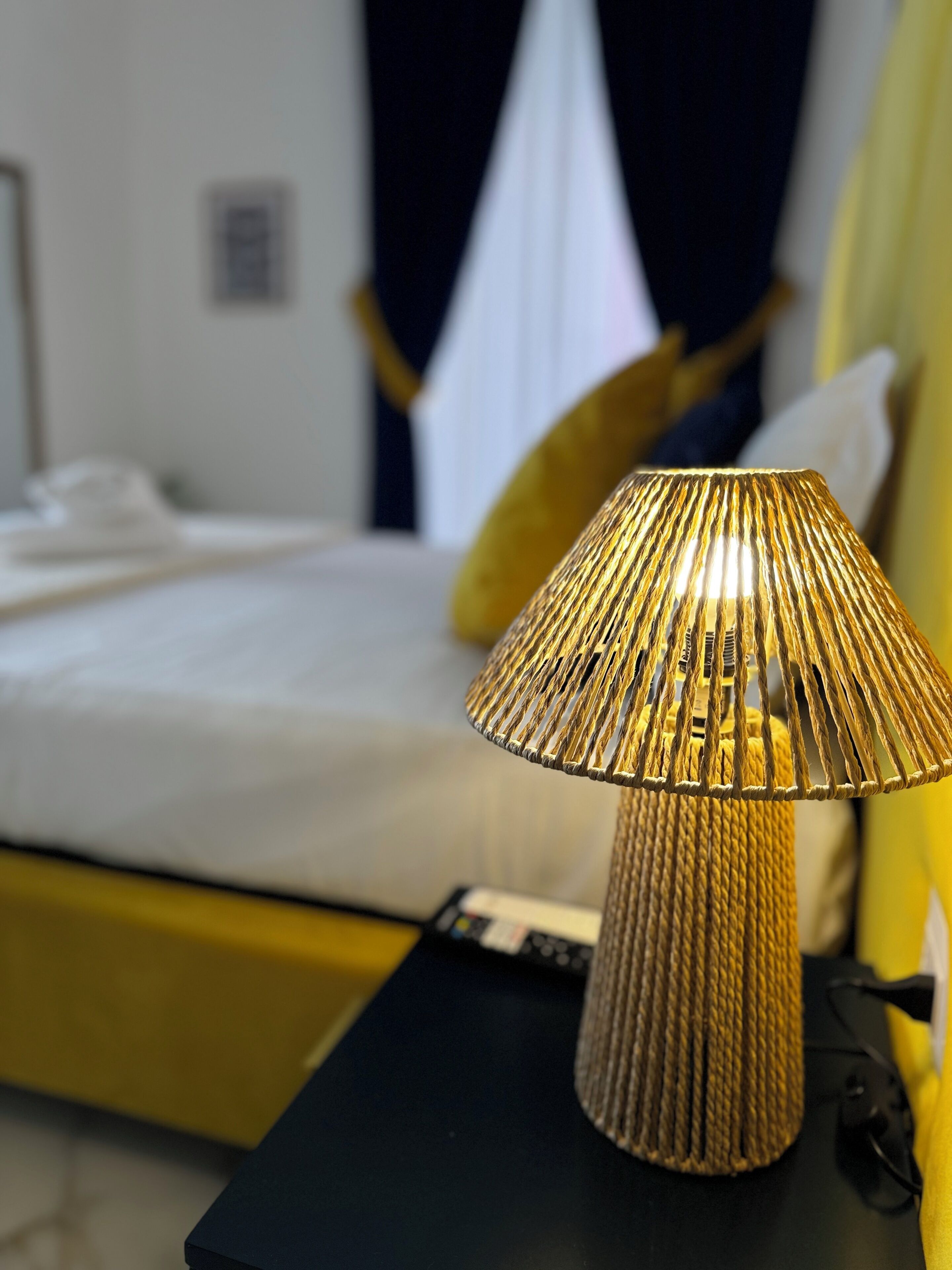 A bed with a white bedspread and a yellow pillow, a black nightstand with a lamp, and a yellow wall.