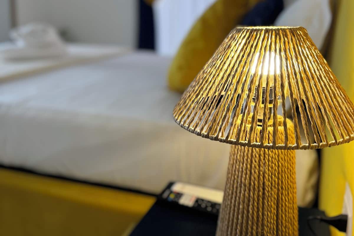 Un lit recouvert d’une couvre-lit blanche et orné d’un coussin jaune, une table de chevet noire avec une lampe, et un mur jaune.