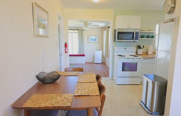 A compact kitchen with a microwave, stove, and refrigerator. A dining table with woven placemats and a black bowl is in the foreground.