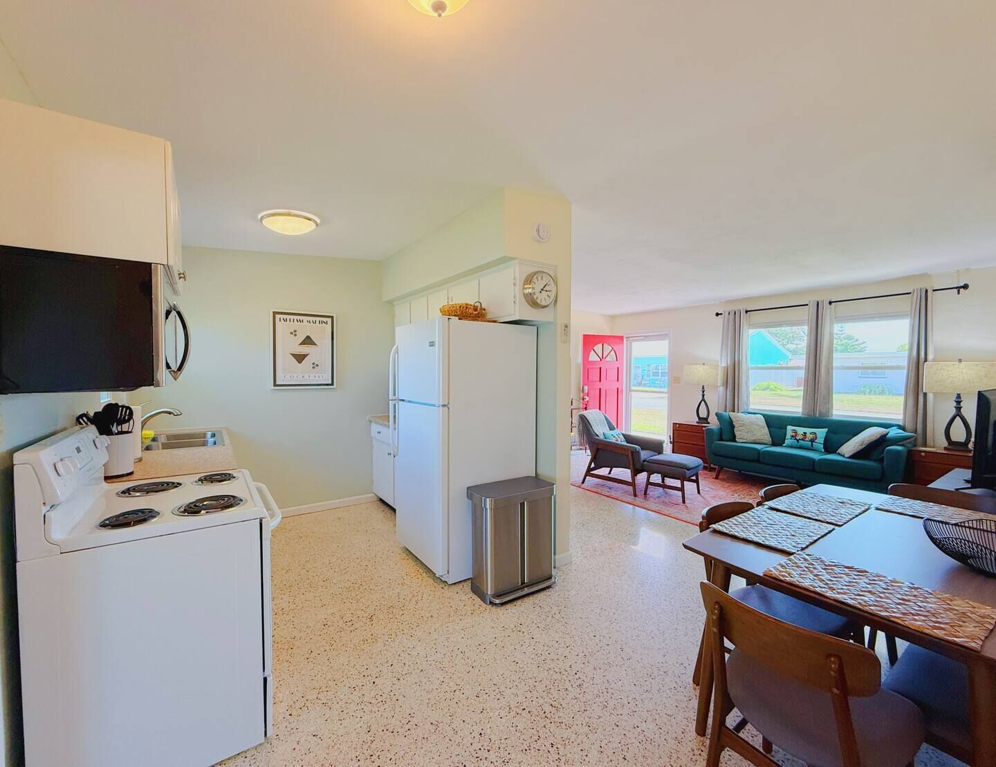 A compact kitchen with a stove, refrigerator, and sink. There is a dining area with a table and chairs, and a living room with a couch and a window.