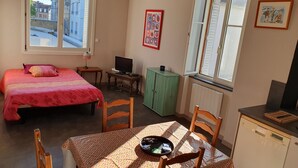 Ein kleines, helles Zimmer mit einem Bett, einem Schreibtisch mit Computer, einem Esstisch mit Stühlen und Blick auf die Straße durch ein Fenster.